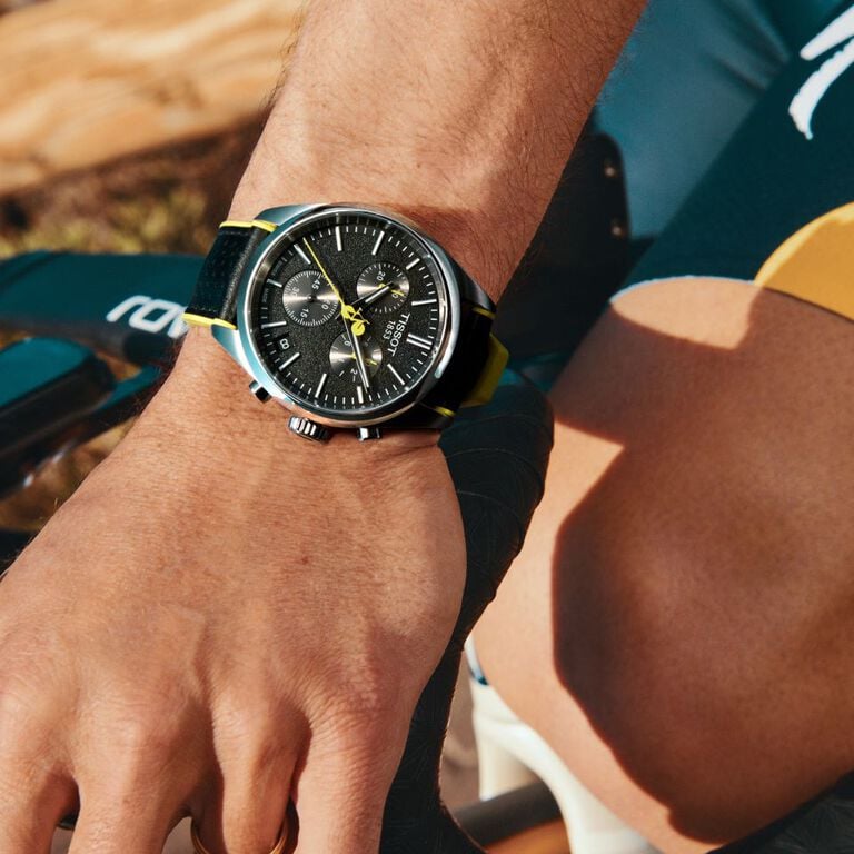 Siyah kadranlı, sarı detaylı ve uyumlu kayışlı Tissot PR 100 Tour de France saat takan bir erkek bileğinin bisiklet bağlamındaki yakın plan görüntüsü.