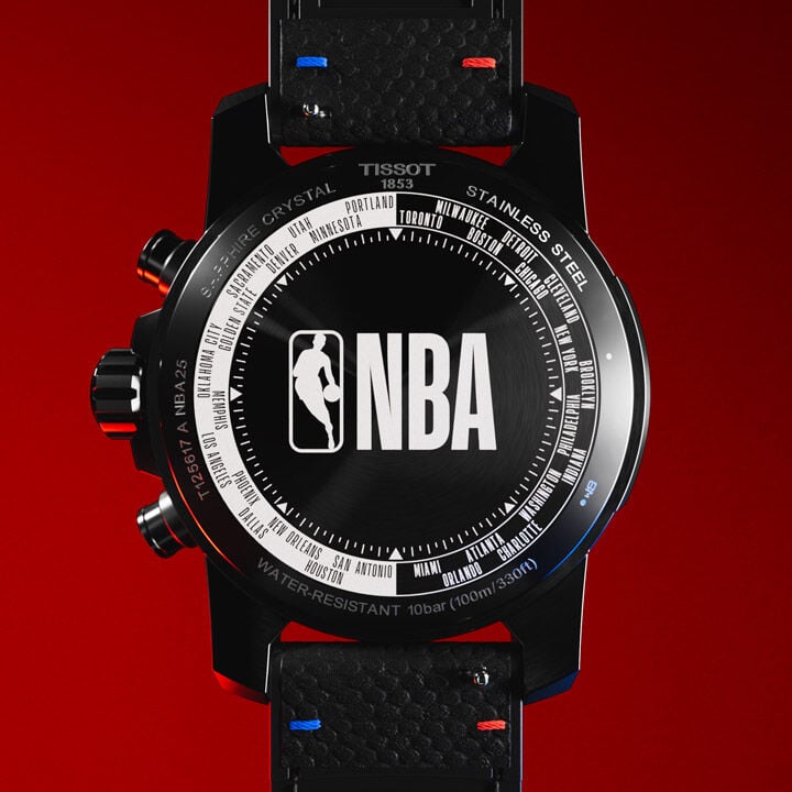 Close-up of the case back of the Tissot Supersport Chrono NBA Edition watch, featuring a bold NBA logo in the center, surrounded by the names of all NBA teams engraved in a circular pattern. The black stainless steel case is contrasted by red and blue stitching on the leather strap, set against a striking red background.